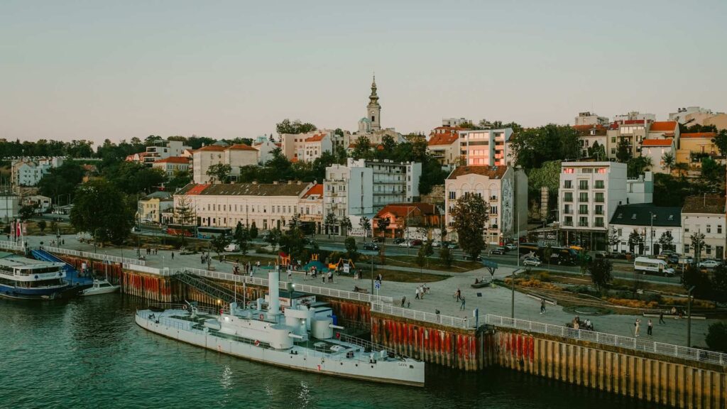 View of Belgrade's riverfront with boats docked, historic buildings, and a church steeple rising in the background.