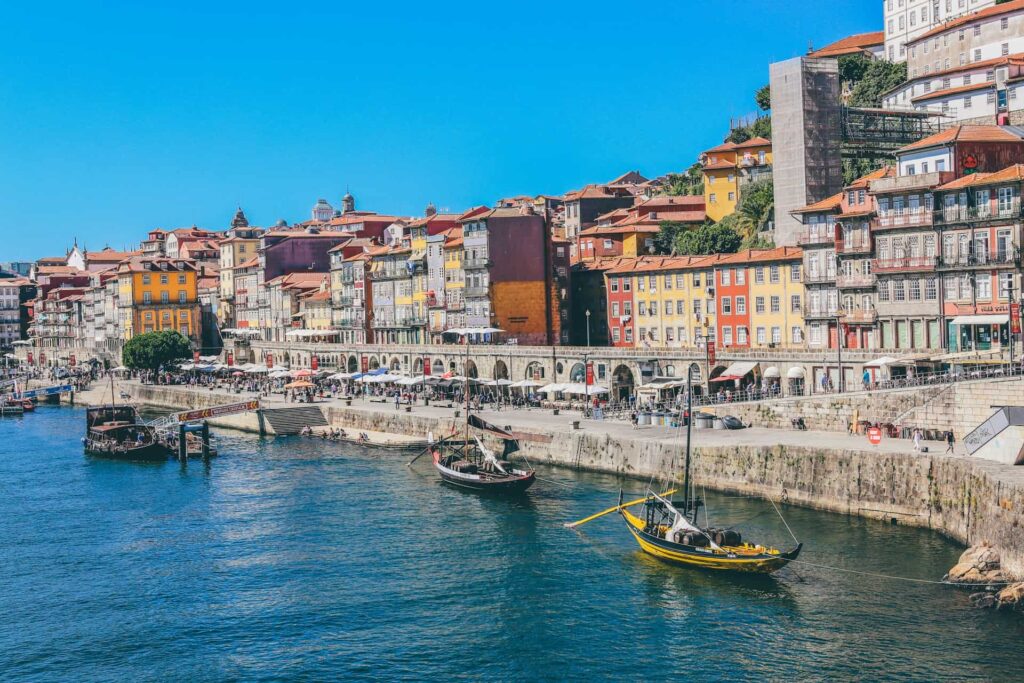 Colourful waterfront buildings in Porto with traditional boats moored on the Douro River and crowds along the promenade.